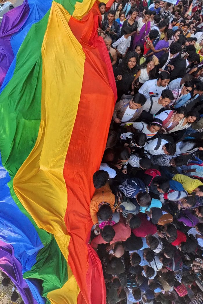 Aerial view of a lively LGBTQ pride parade in Kolkata, West Bengal, with a large rainbow flag and a diverse crowd.